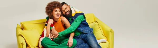 African American friends in colorful attire sitting together on a yellow chair against a grey background, showcasing friendship. — Stock Photo