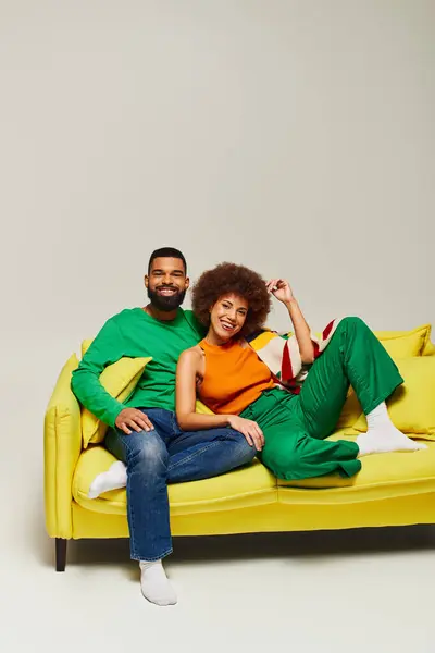Happy African American friends in vibrant clothes sitting on a yellow couch against a grey background. — Stock Photo