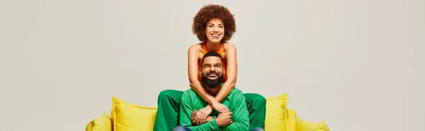 A man in vibrant attire sits atop a woman on a yellow couch, showcasing the bond of friendship in colorful contrast. — Stock Photo