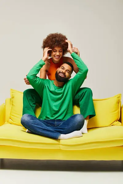 Homme et femme, amis afro-américains, assis joyeusement sur un canapé jaune en tenue vibrante, sur fond gris. — Photo de stock