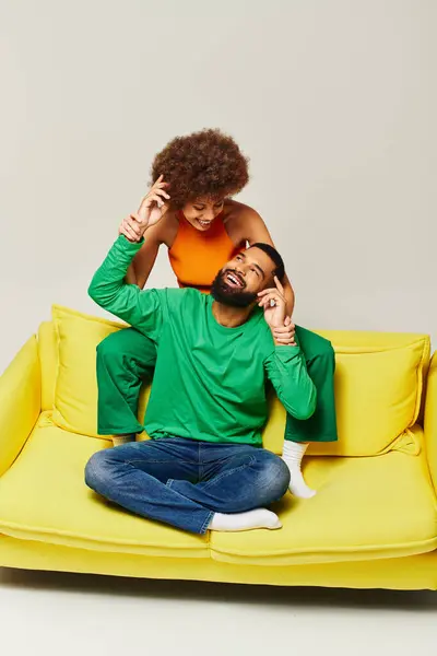 Un homme et une femme afro-américains sont heureux de s'asseoir sur un canapé jaune en vêtements vibrants sur un fond gris. — Photo de stock