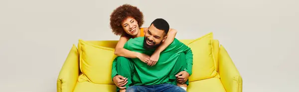 Ein fröhlicher afroamerikanischer Mann und eine fröhliche afroamerikanische Frau sitzen in lebendigen Kleidern auf einem gelben Stuhl vor grauem Hintergrund.. — Stockfoto