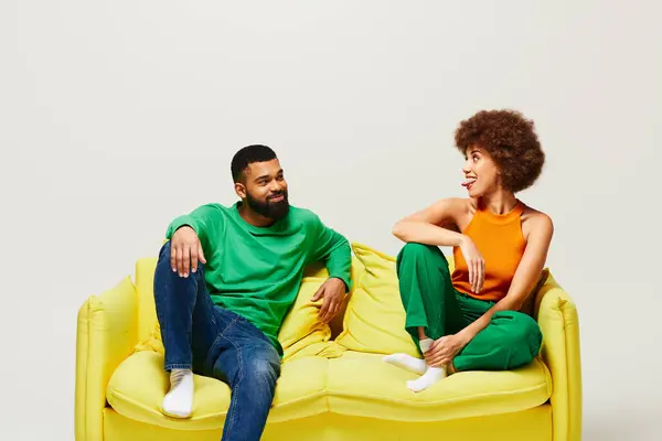 Heureux amis afro-américains en vêtements vibrants assis ensemble sur un canapé jaune sur un fond gris. — Photo de stock