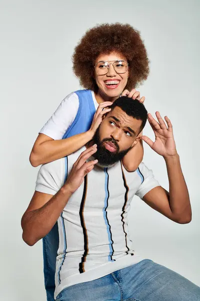 Um homem afro-americano elegante brinca sentado em cima de uma cabeça de mulher contra um fundo cinza, mostrando amizade e confiança. — Fotografia de Stock