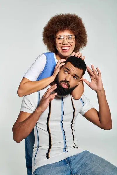 A stylish African American man playfully sits on top of a womans head against a grey background, showcasing friendship and trust. — Stock Photo