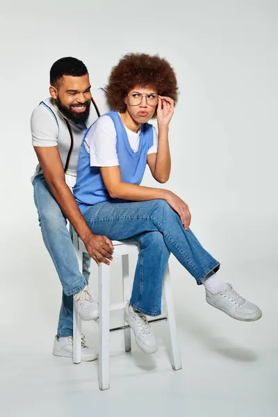 An African American man and a woman, dressed stylishly, sharing a stool in a pose of friendship on a grey backdrop. — Stock Photo
