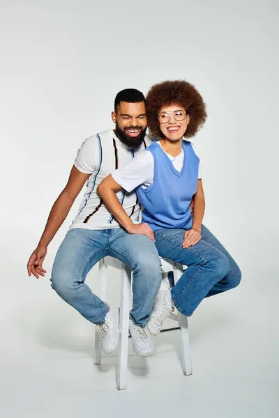 Amis afro-américains dans des vêtements élégants, mettant en valeur l'amitié à travers une pose tout en étant assis sur une chaise sur un fond gris. — Photo de stock