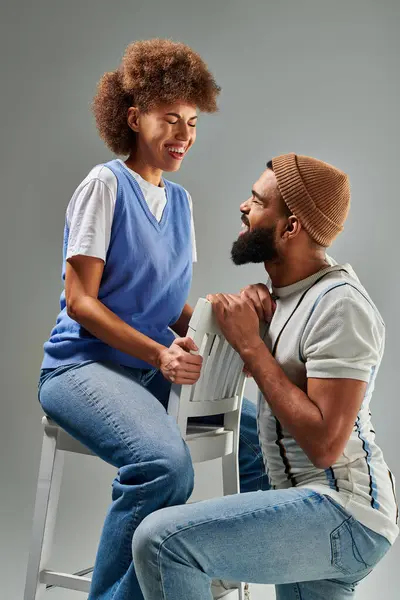 Un homme et une femme afro-américains vêtus élégamment, assis sur des tabourets sur un fond gris, respirant un air d'amitié. — Photo de stock