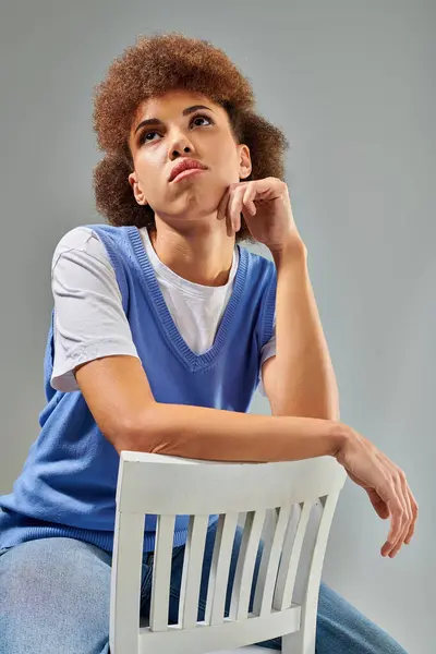 Une femme en tenue élégante s'assoit sur une chaise blanche, expression réfléchie avec la main sur le menton, sur un fond gris. — Photo de stock