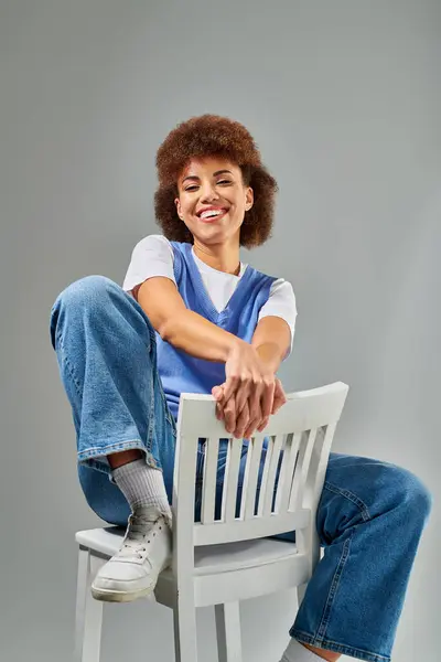 A stylish African American woman in chic attire sits elegantly on top of a white chair against a grey background. — Stock Photo