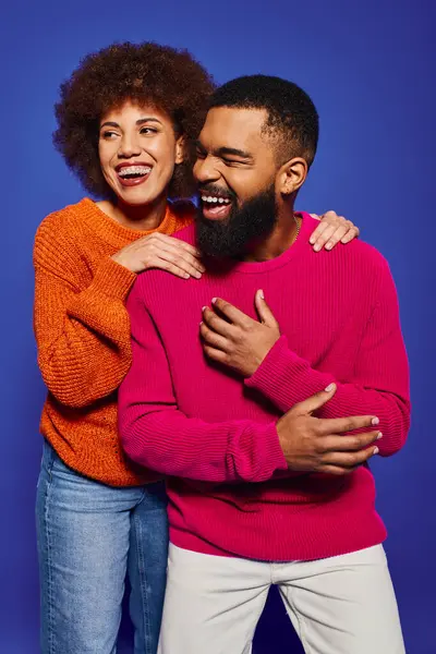 Ein junger afroamerikanischer Mann und eine junge afroamerikanische Frau, Freunde, in lebhafter, lässiger Kleidung, die eine innige Umarmung auf blauem Hintergrund teilen. — Stockfoto