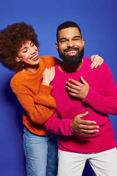 A young African American man and woman, friends posing playfully in vibrant casual attire on a blue background. — Stock Photo