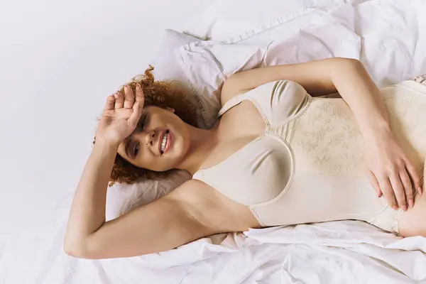 A young, curvy redhead woman reclining on a bed in a white bodysuit, against a grey background. — Foto stock