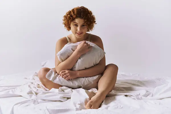 A young, curvy redhead woman in lingerie sits on a bed surrounded by pillows. — Foto stock