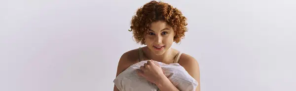 A young curvy redhead woman in lingerie delicately holds a fluffy pillow in her hands on a grey background. — Stockfoto