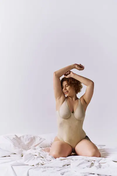 Young, curvy redhead woman in bodysuit sitting gracefully on bed against grey backdrop. — Stockfoto