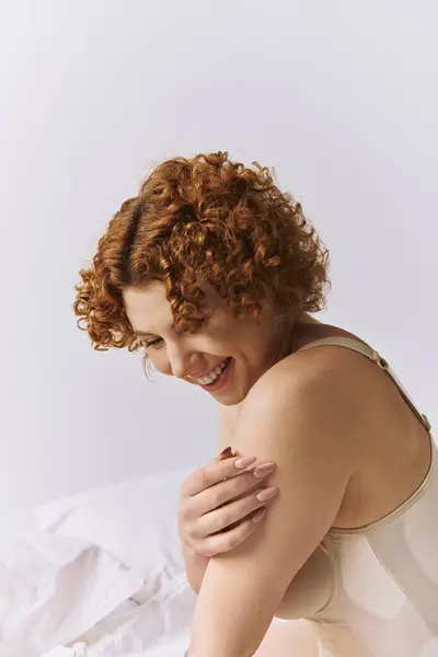 A young, curvy redhead woman in lingerie sits on a bed, laughing cheerfully. — Stock Photo