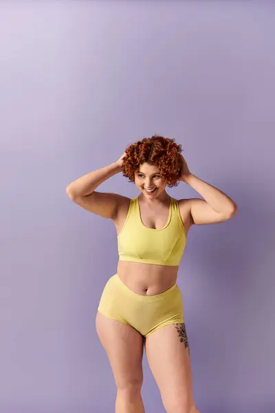 A young curvy redhead woman in a yellow bikini striking a pose against a bold purple backdrop. — стоковое фото