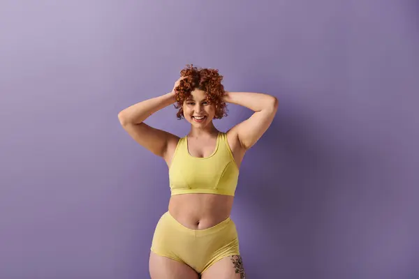 A young, curvy redhead woman confidently poses in a yellow bikini against a striking purple background. — Stock Photo