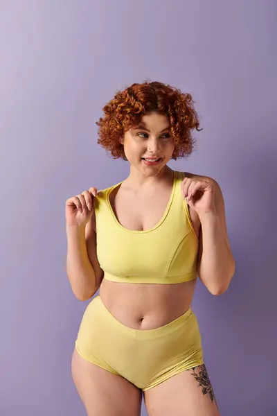 Young curvy redhead woman showcasing strength in yellow sports bra against purple backdrop. — Stock Photo