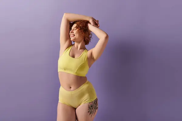 A young, curvy redhead woman in a yellow bikini striking a pose on a purple background. — Stock Photo