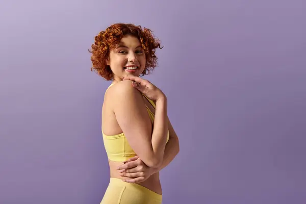 Curvy young redhead in yellow lingerie striking a pose against a purple backdrop. — Photo de stock
