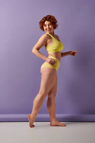 A young, curvy redhead woman in a yellow bikini striking a pose against a bold purple wall. — Stock Photo