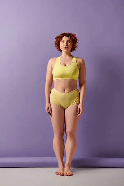 A young, curvy redhead woman in a yellow bikini stands confidently in front of a vibrant purple wall. — Stockfoto