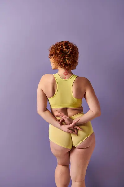 Young redhead woman flaunts curves in yellow bikini against vibrant purple backdrop. — Stock Photo