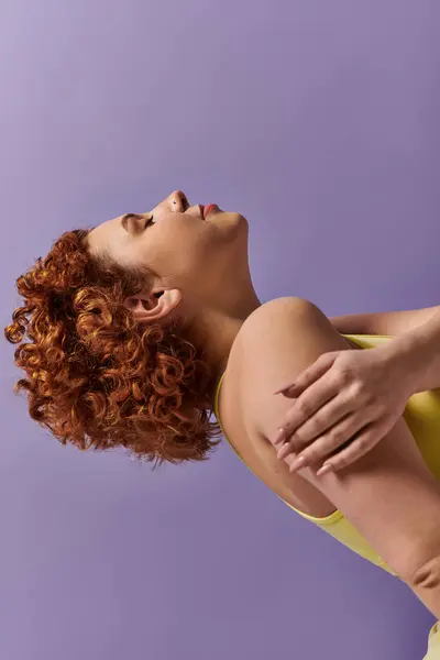 A curvy young redhead woman, dressed in a yellow shirt, performs a back stretch against a purple background. — Stock Photo