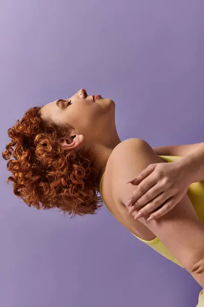 A curvy young redhead woman, dressed in a yellow shirt, performs a back stretch against a purple background. — стоковое фото