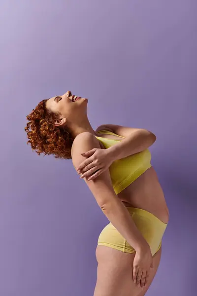 Curvy redhead woman in yellow bikini poses against vibrant purple backdrop. — Stockfoto