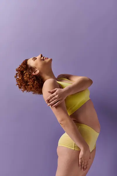 Curvy redhead woman in yellow bikini poses against vibrant purple backdrop. — Stock Photo