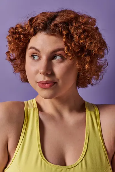 A young, curvy redhead woman stands confidently in a yellow tank top against a vibrant purple background. — Stock Photo