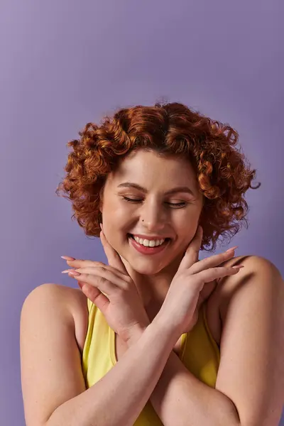 A young, curvy redhead woman in yellow underwear strikes a playful pose, hands on face, against a purple background. - foto de stock
