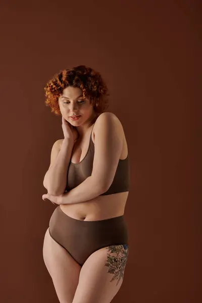A curvy redhead woman strikes a pose in a brown bikini against a matching background. - foto de stock