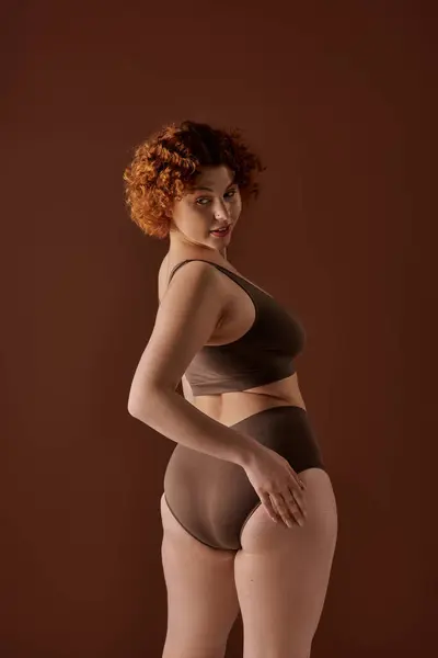 Young, curvy redhead woman in a brown bikini striking a pose on a brown background. — Stock Photo