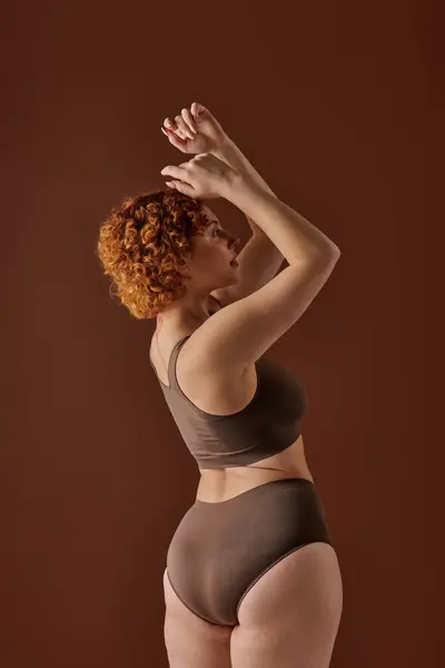 Young curvy redhead woman in brown bikini striking a pose with hands raised. — Stock Photo