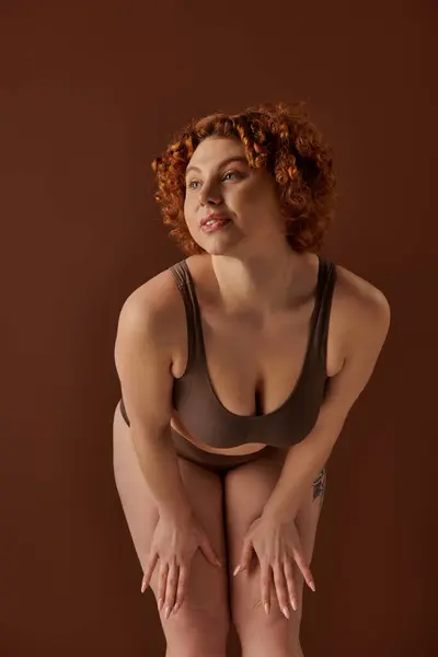 Curvy redhead woman in underwear posing on a brown backdrop. — Stockfoto