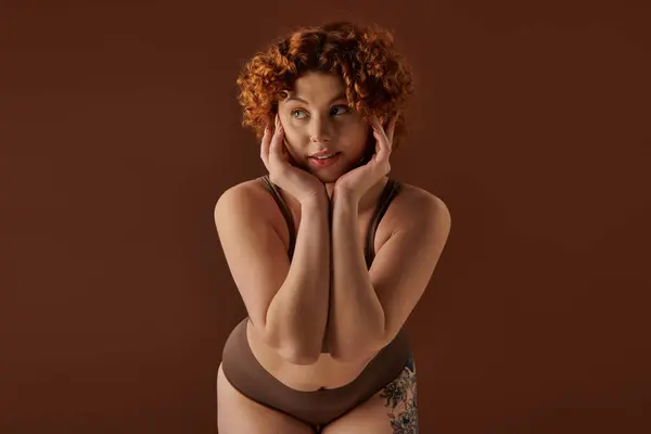 A curvy redhead woman showcases elegance and sensuality in lingerie against a brown background. — стокове фото
