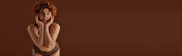 A young, curvy redhead woman exudes confidence in a bikini, striking a pose on a plain brown background. — стоковое фото