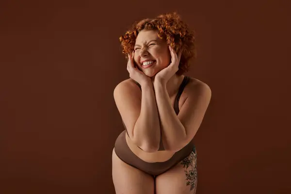 A young, curvy redhead woman poses seductively in a bikini against a warm brown backdrop. — стокове фото
