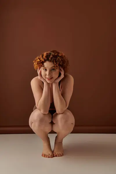 A young, curvy redhead woman crouches gracefully on the floor in her underwear against a brown backdrop. — Stock Photo