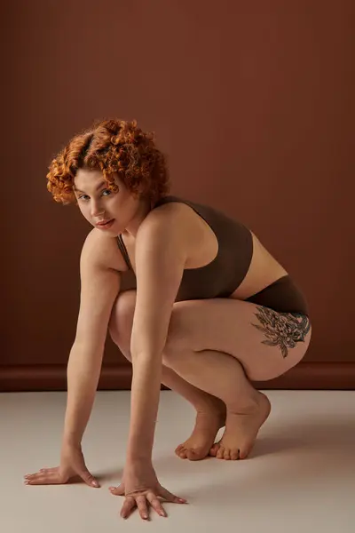 A crouching young, curvy woman with fiery red hair on a brown background. — Stock Photo