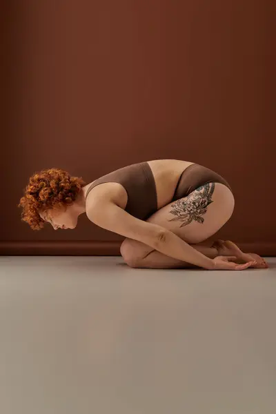 A curvy redhead crouches on the floor displaying her vibrant tattoos. - foto de stock
