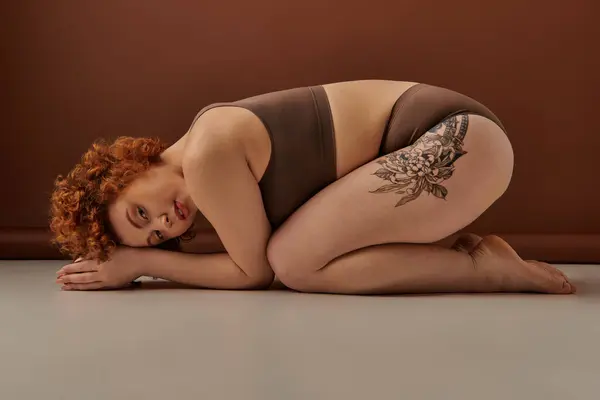 A curvy, young redhead woman in underwear crouches on the floor, showcasing her tattoos. — стоковое фото
