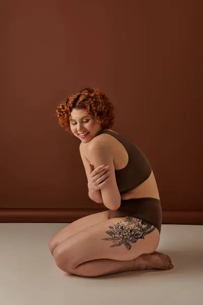 A young, curvy redhead woman sits gracefully on the floor in a cozy brown room. — стокове фото