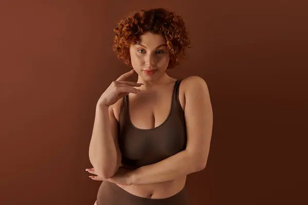 Young curvy redhead woman exuding confidence in brown bra on soft brown backdrop. — Stockfoto