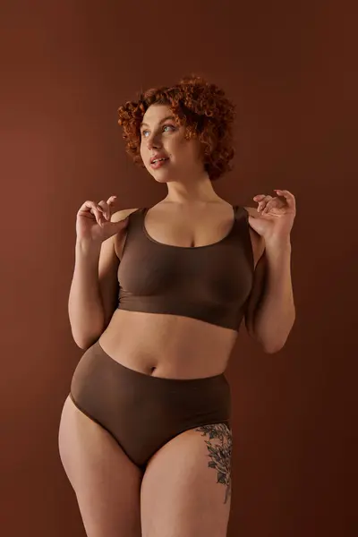 A young and curvy redhead woman in a brown bikini poses gracefully on a warm-toned brown background. — Foto stock