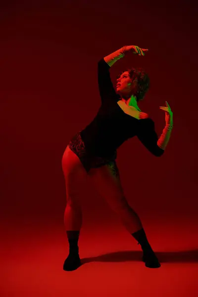 A young curvy redhead woman in a bodysuit dances gracefully in front of a vivid red background. — стоковое фото